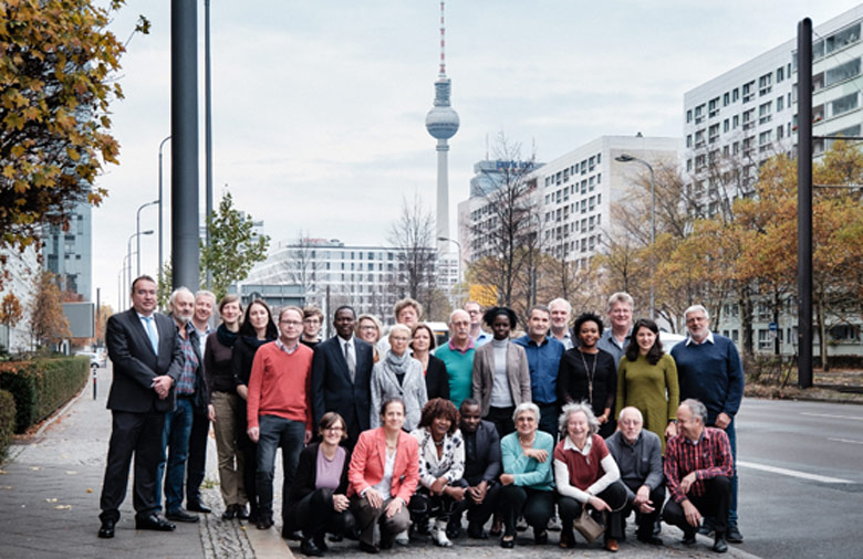 Rund dreißig Teilnehmende haben sich an einer Straße zu einem Gruppenbild zusammengestellt und lächeln in Richtung des Fotografen. Im Hintergrund ist der Berliner Fernsehturm zu erkennen.